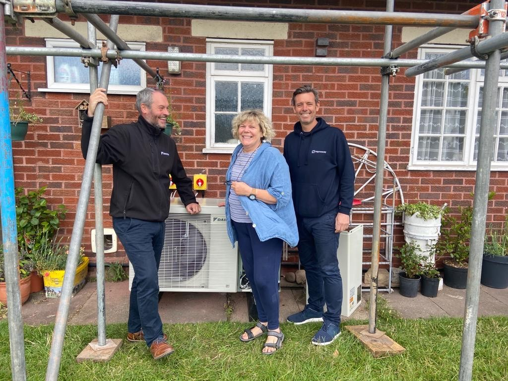 two installers and one customer stand smiling in front of a new air source heat pump in a domestic garden