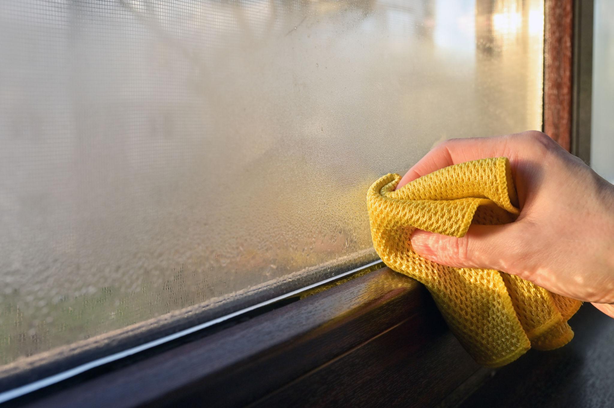 Hand wiping condensation from a window, showing moisture build-up inside the home