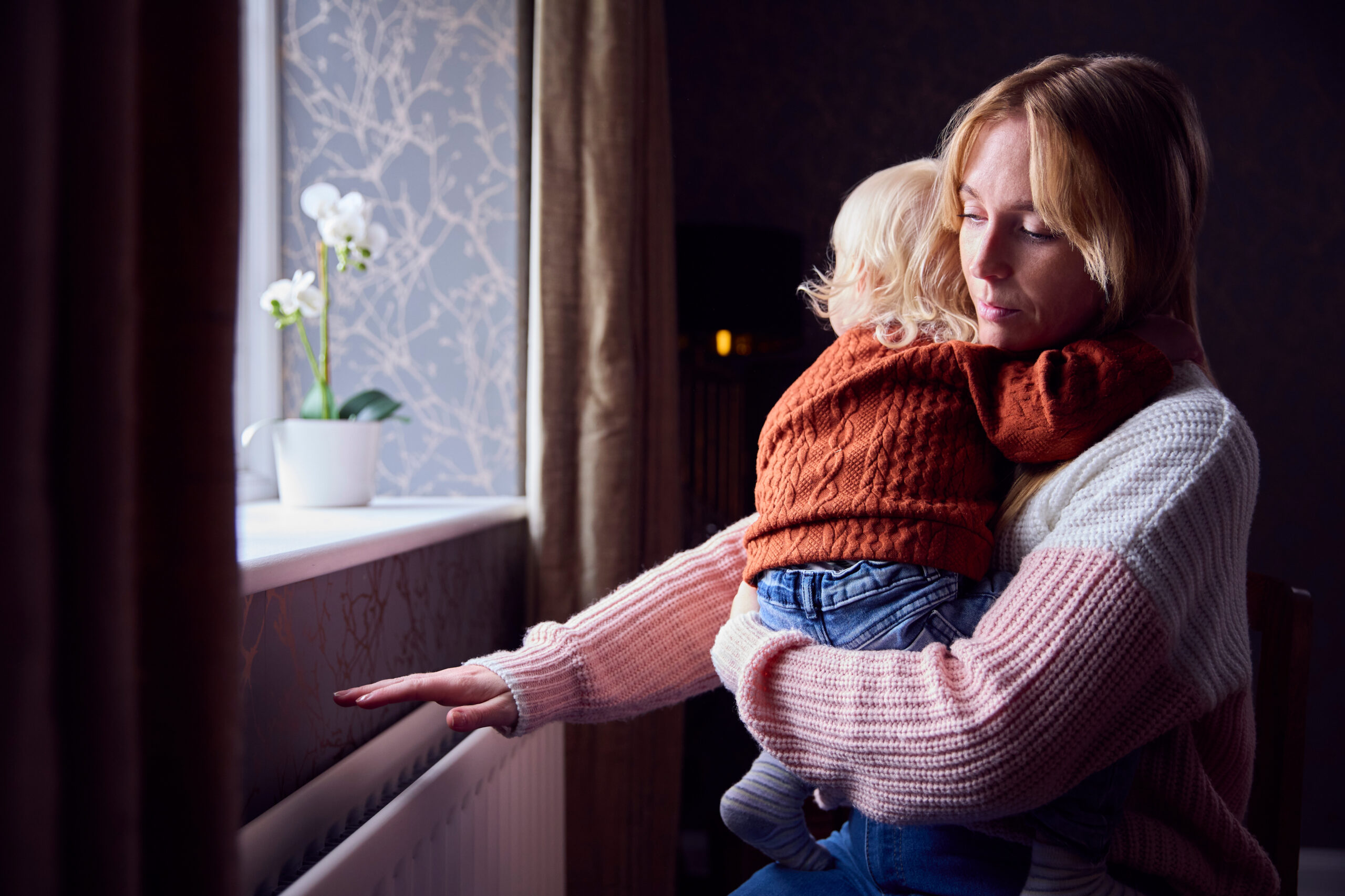 A blonde woman in a pink and white sweater holds a young child in a rust-orange knit sweater. She looks down thoughtfully while reaching out to touch a white radiator beneath a window with a potted white orchid.