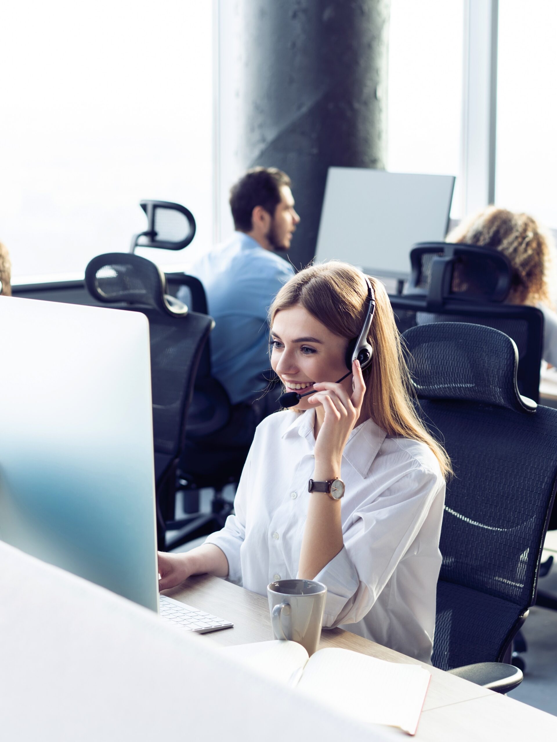 A smiling female customer service representative wearing a headset sits at her desk in a modern office, looking at a computer screen. Other colleagues are visible working in the background.