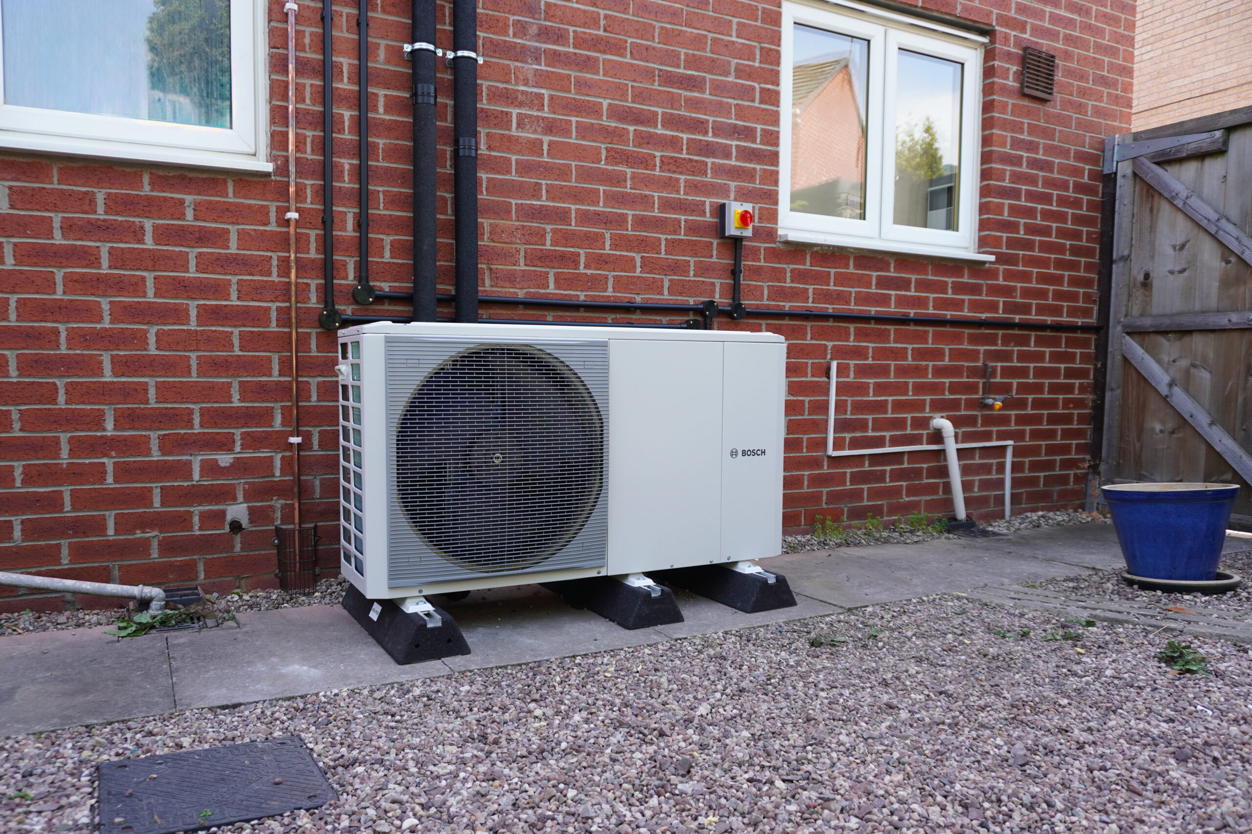A white Bosch air source heat pump installed outdoors against a red brick wall, connected to insulated black and copper pipework.