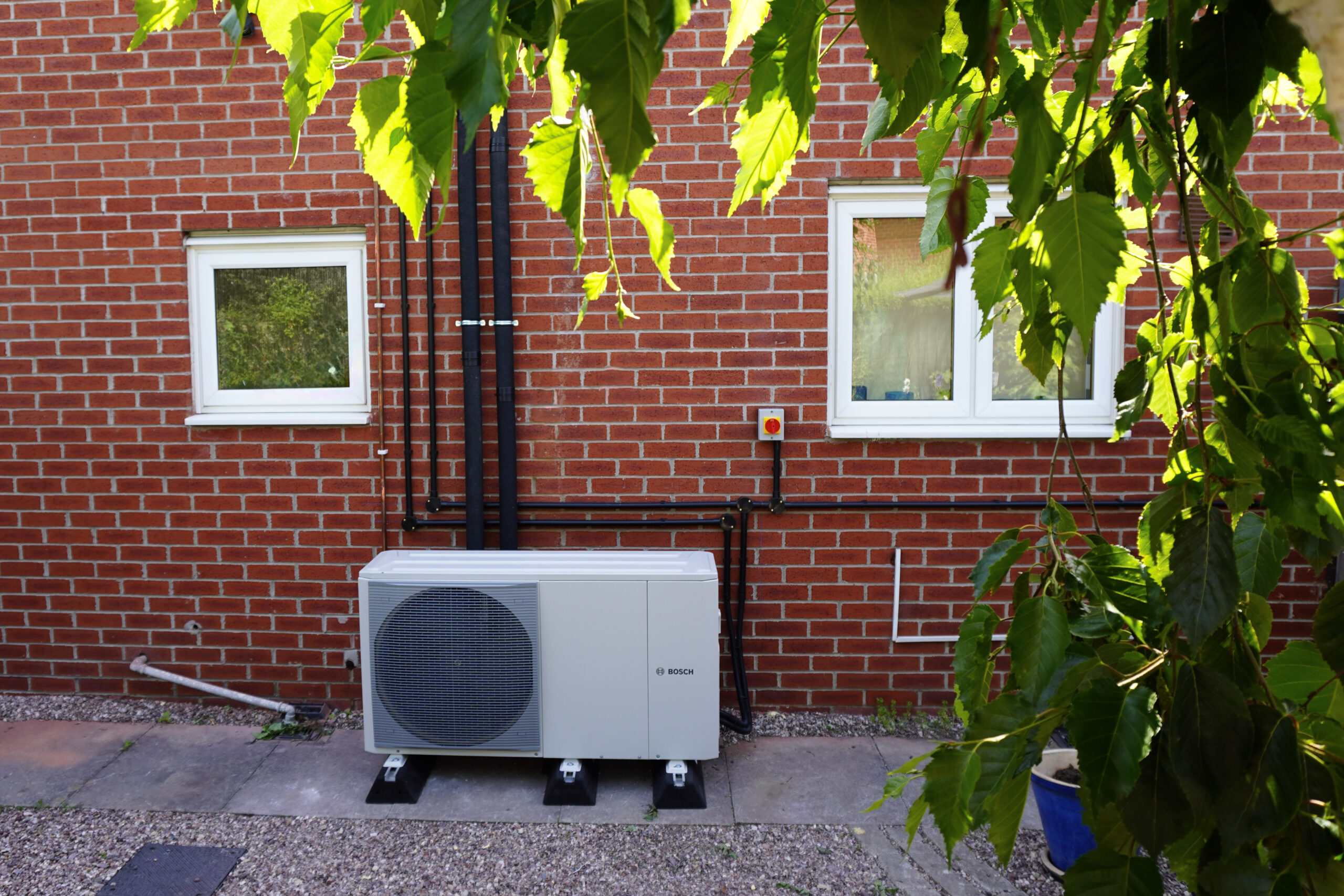 A white Bosch air source heat pump installed outside against a red brick wall, situated between two white-framed windows with green leaves framing the top and right side of the image.