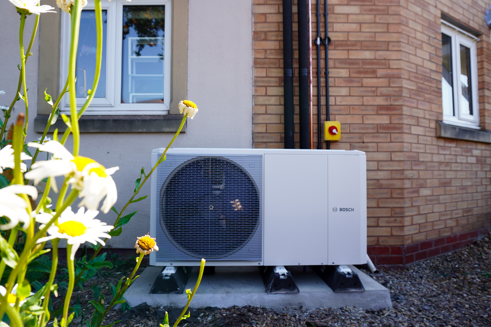Close-up of a Bosch outdoor heat pump unit mounted outside a house, framed by white flowers in the foreground.