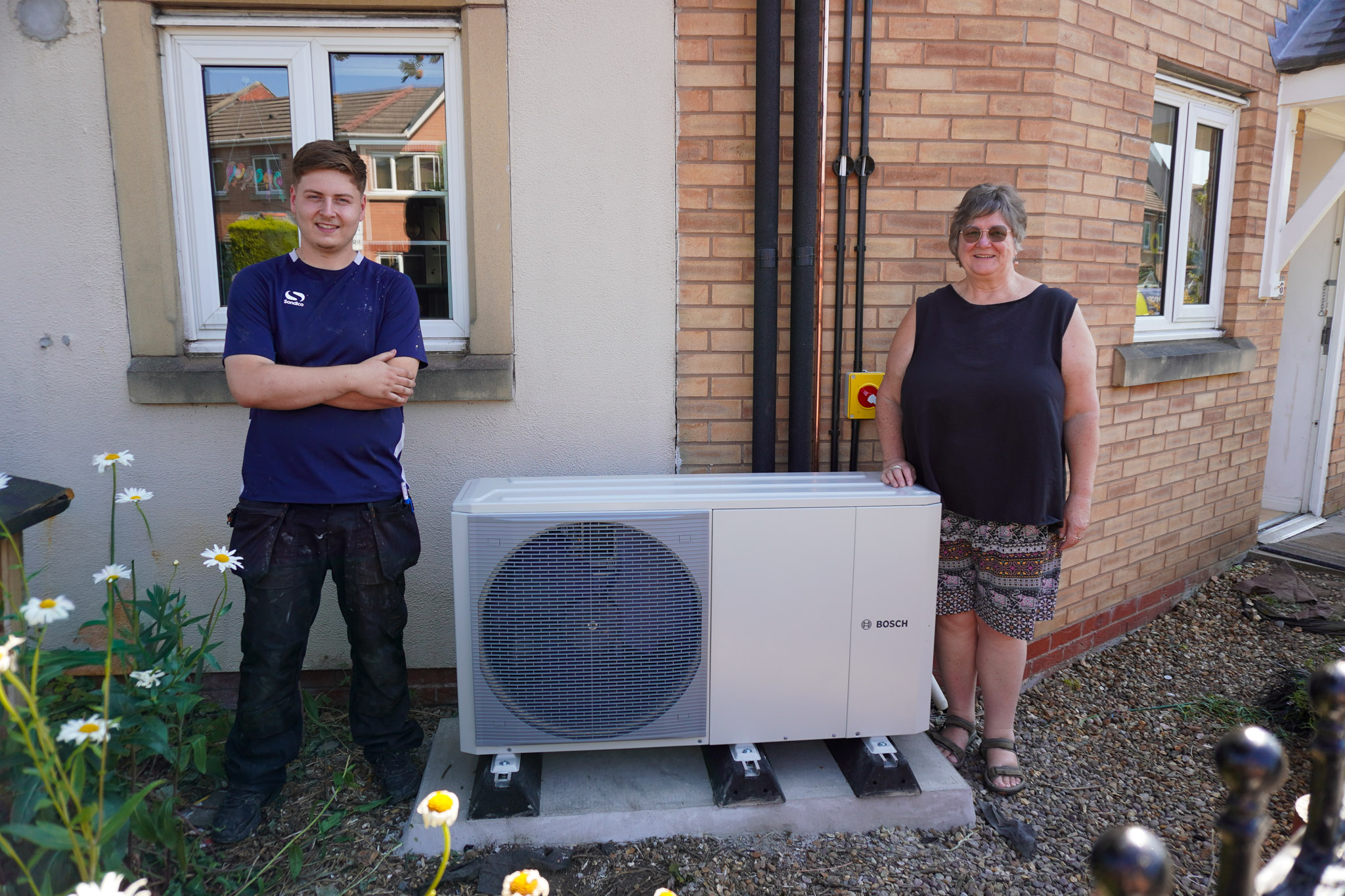 An Improveasy engineer and homeowner Karen standing outside her home next to a newly installed Bosch air source heat pump unit.