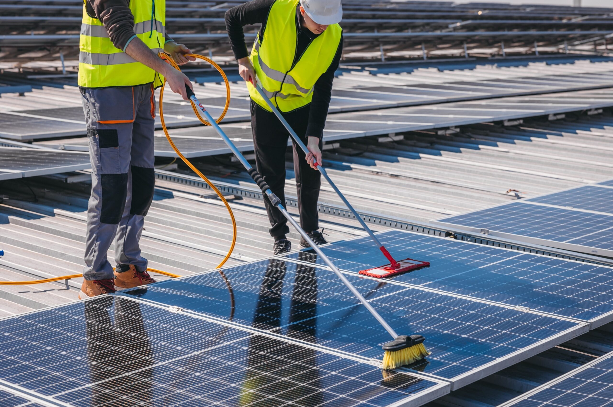 Two technicians in high-visibility vests using long telescoping brushes to clean dirty rooftop solar panels.