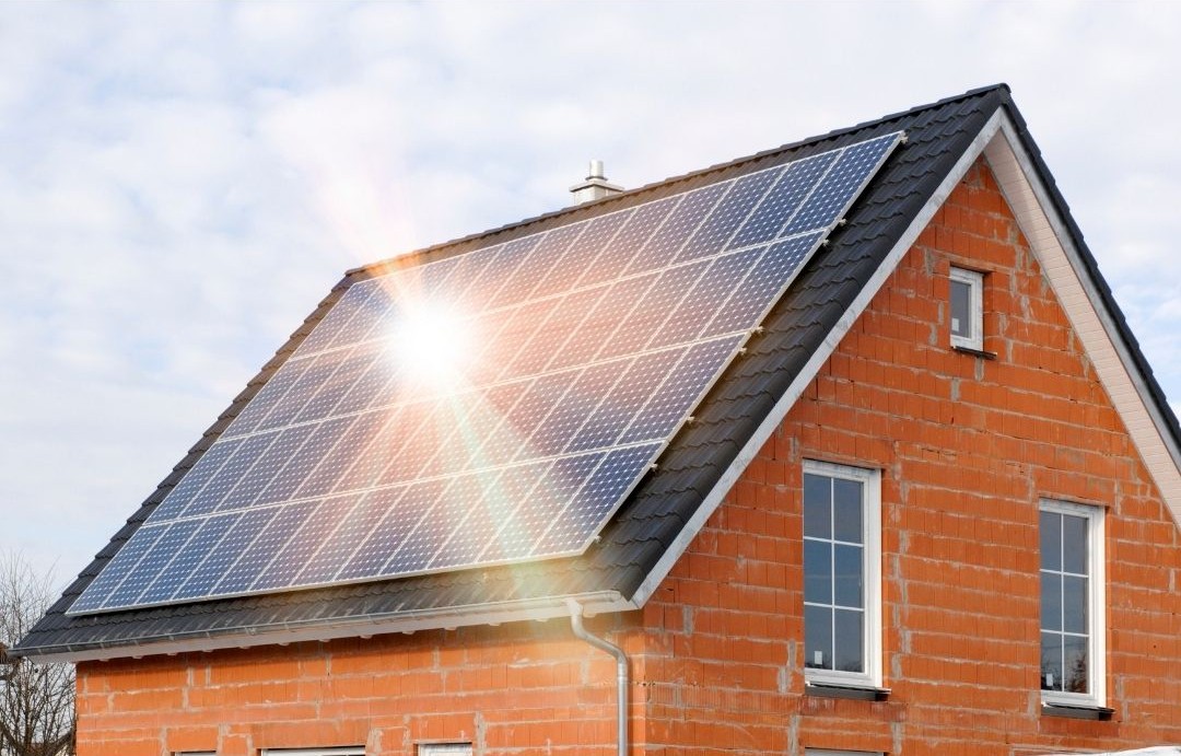 Bright sunlight gleaming off a large solar panel system installed on the tiled roof of a red brick house.