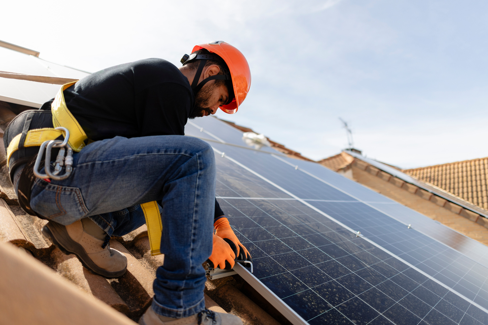 A solar panel technician wearing a hard hat and safety harness crouching on a roof to inspect and secure a panel module.