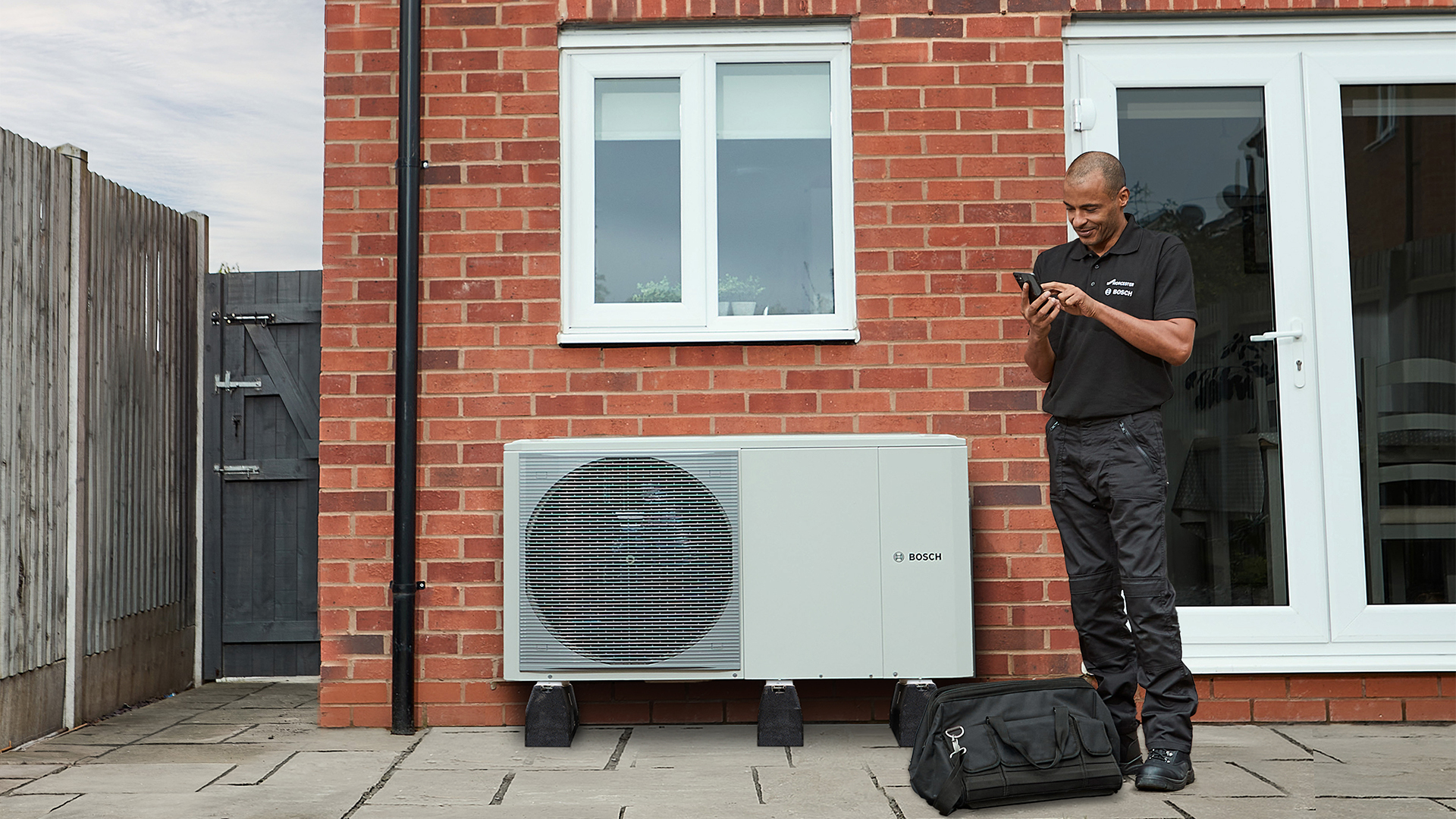 An installer standing next to a Bosch outdoor heat pump unit on a patio, using a smartphone.