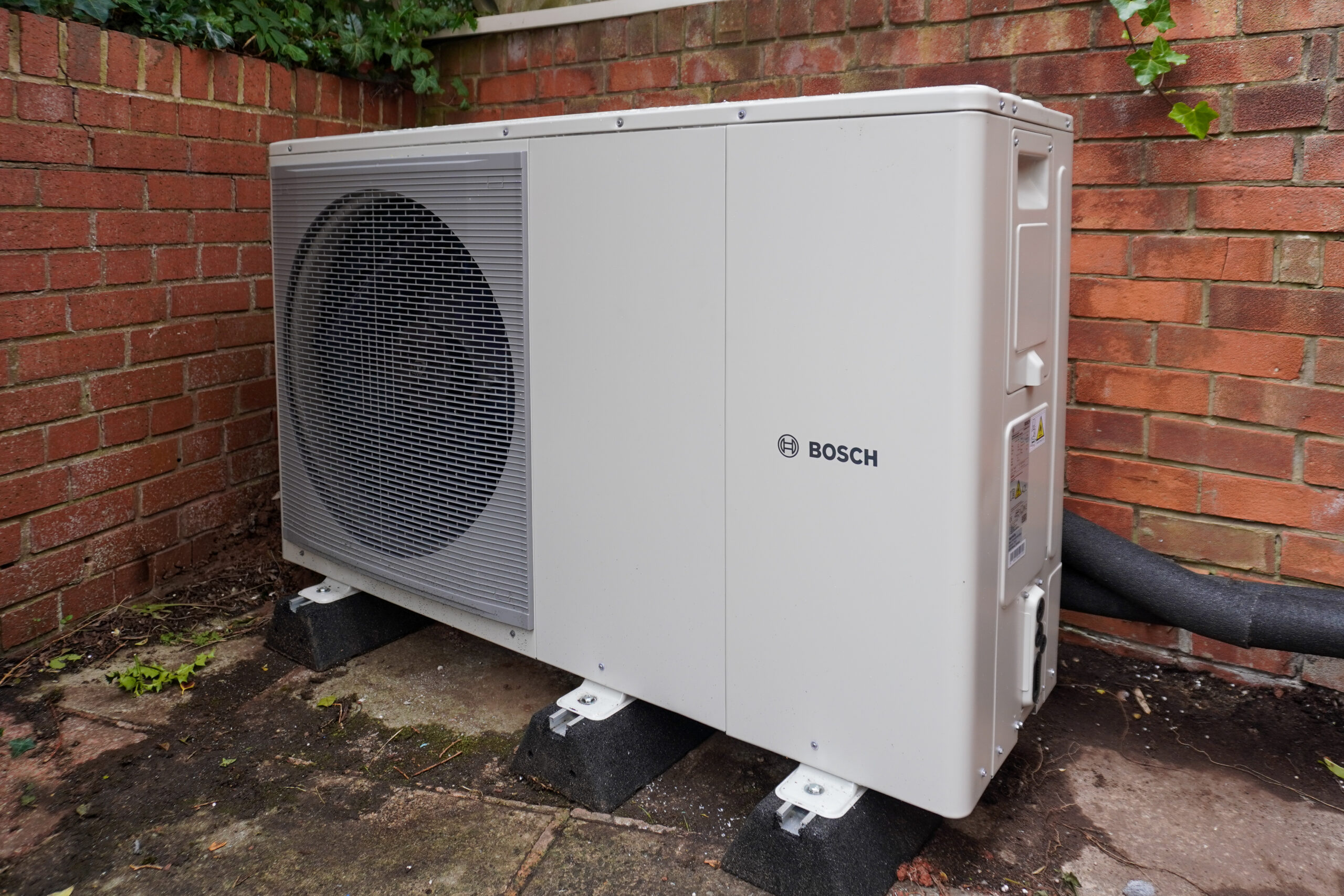 A white Bosch air source heat pump installed outdoors against a red brick wall.