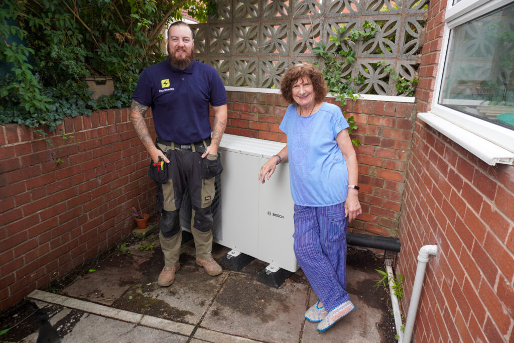 A heating installer and a smiling homeowner standing next to a newly installed Bosch air source heat pump in a garden courtyard
