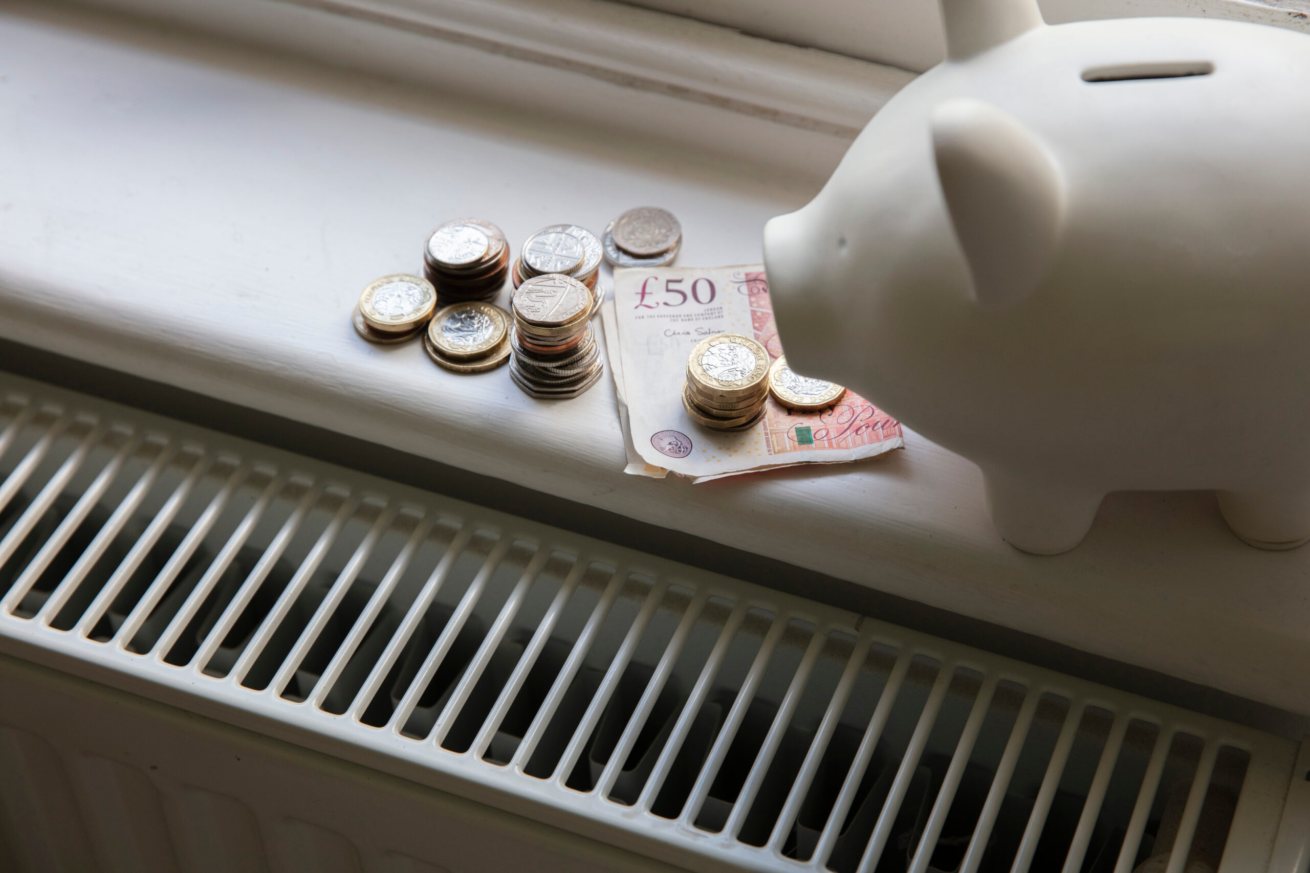 A white ceramic piggy bank sitting on a windowsill next to stacks of British pound coins and a £50 banknote, positioned directly above a home heating radiator.