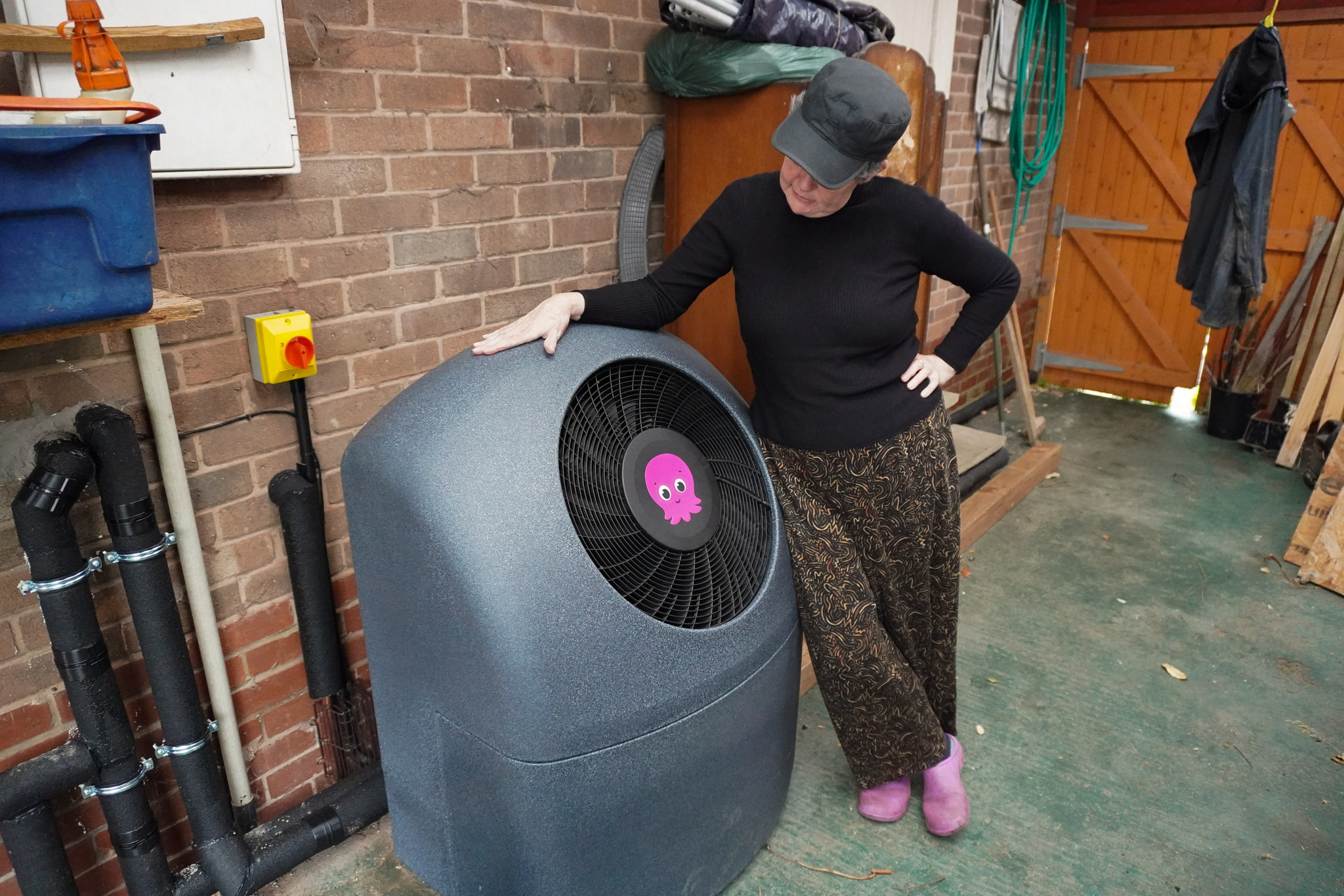 Jane resting her arm on her installed Octopus Cosy heat pump outside her home, looking down at the unit.